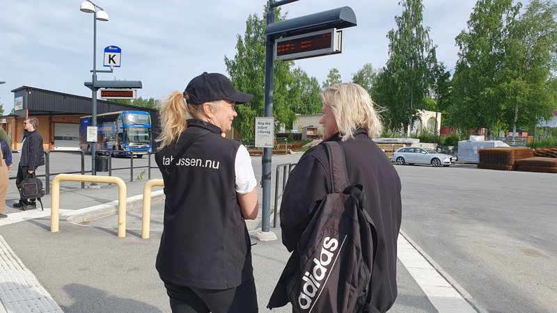 Two people speaking at Skellefteå bus station, digital bus stop signs visible in the background
